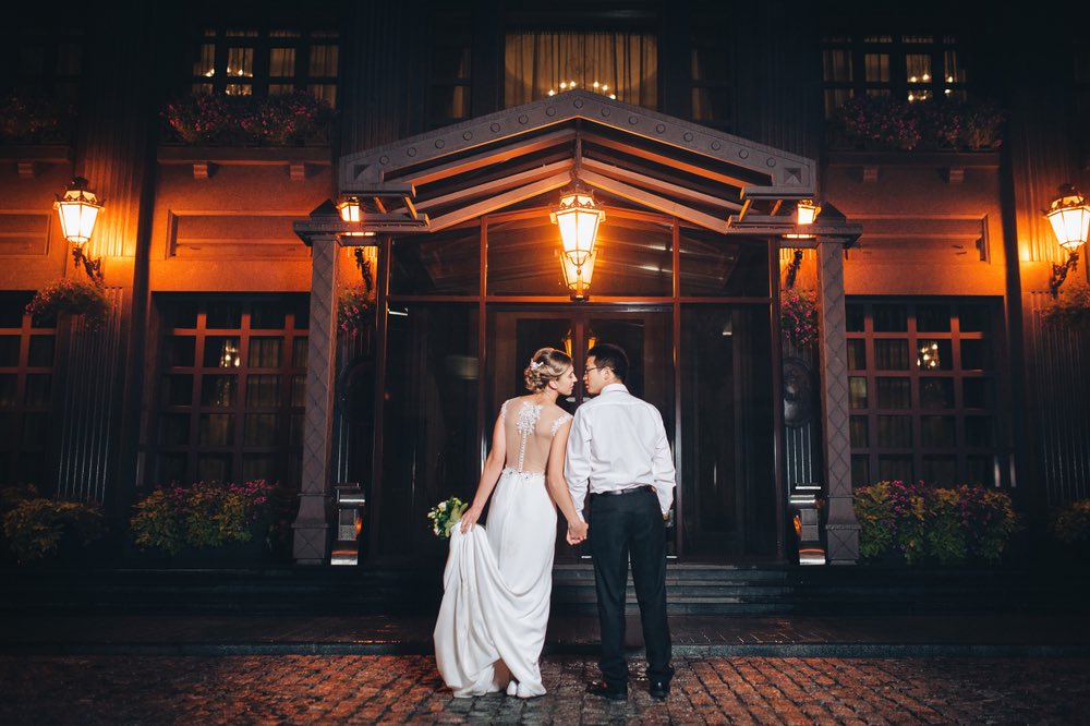 A bride and groom standing on the steps of the entrance to the venue. She is lit up by the lamps above her and the ambient light around her.