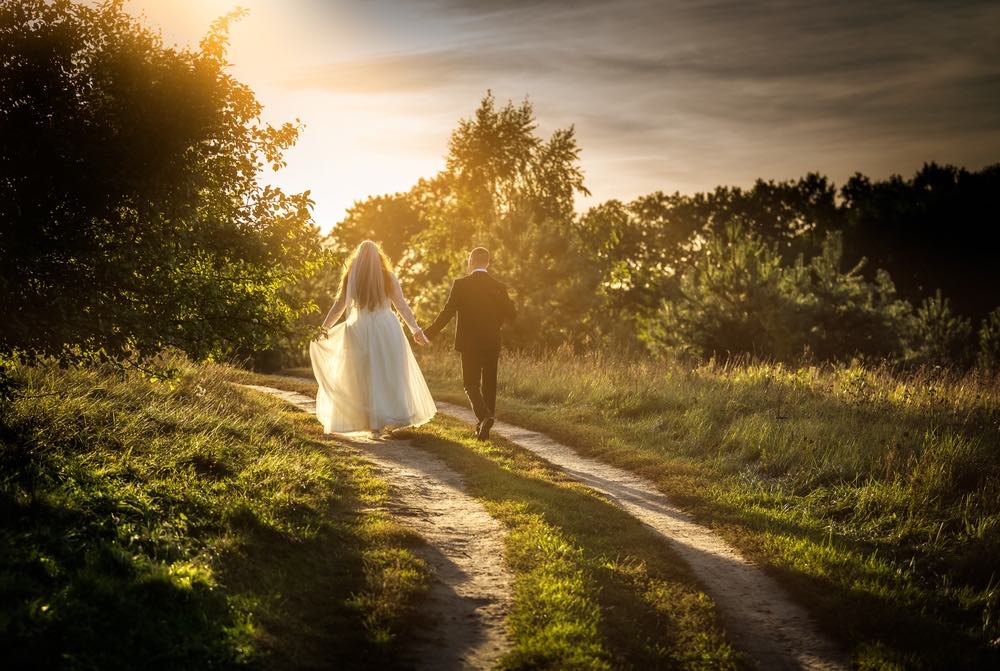 A bride and groom holding hands and walking through a grassy field near sunset.