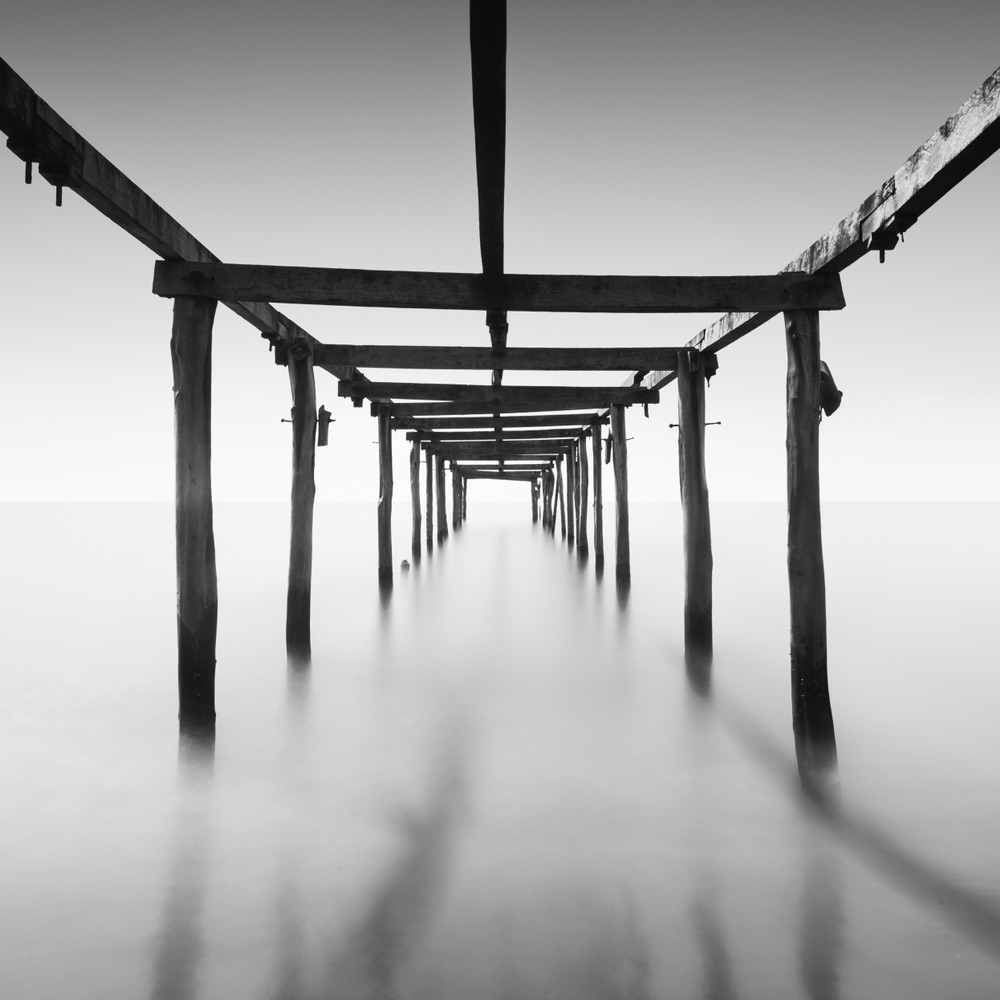 A framework of a dock sits on some water. It's a long exposure B&W so the water is silky smooth. The dock extends into the distance and the shot is from underneath the dock.