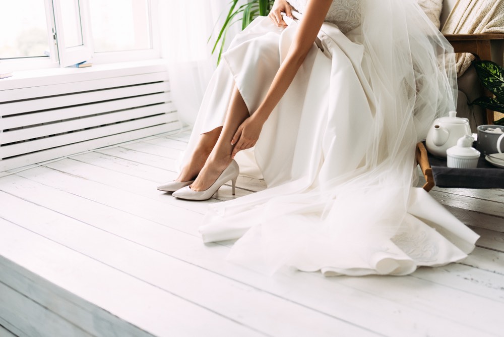 A photo of a bride slipping her shoes on for the wedding/ The dress is draped over the chair in an elegant fashion.