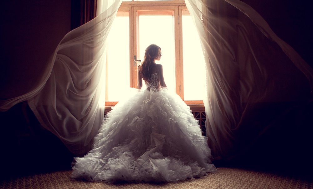 A young bride photographed against an open window within a dark indoor space. An example of tricky lighting conditions in wedding photography which necessitate proper metering.