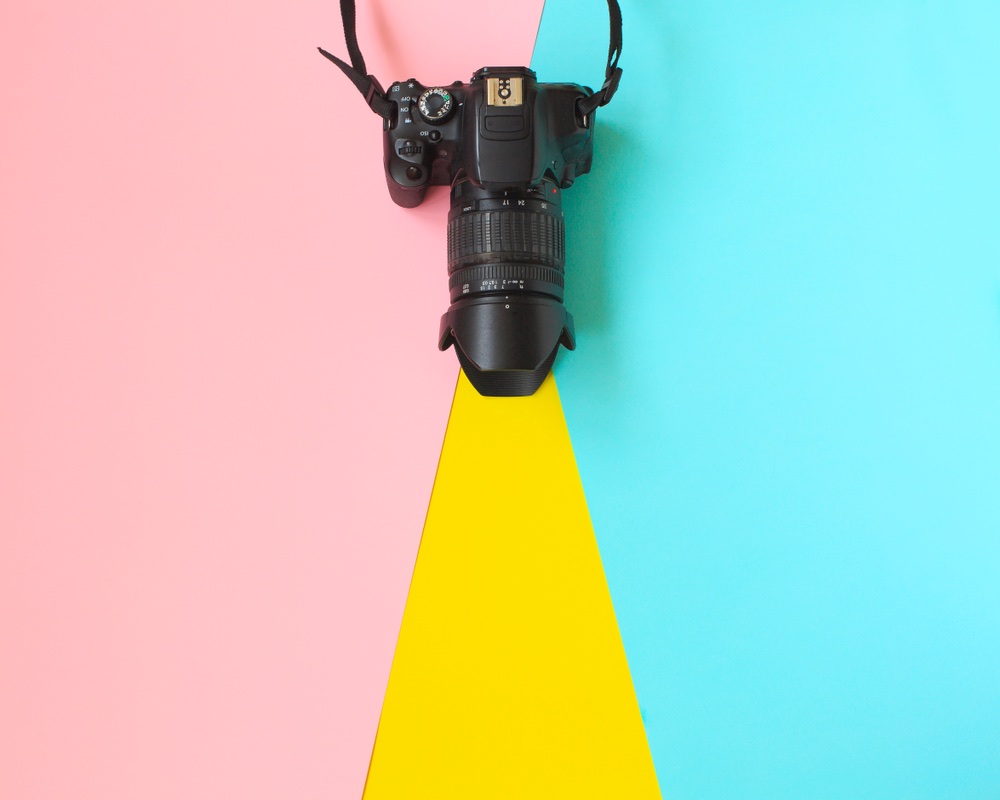 A camera with a zoom lens and lens hood is sitting on a pink, blue, and yellow surface with the lens pointing downward and a minimalist color background.