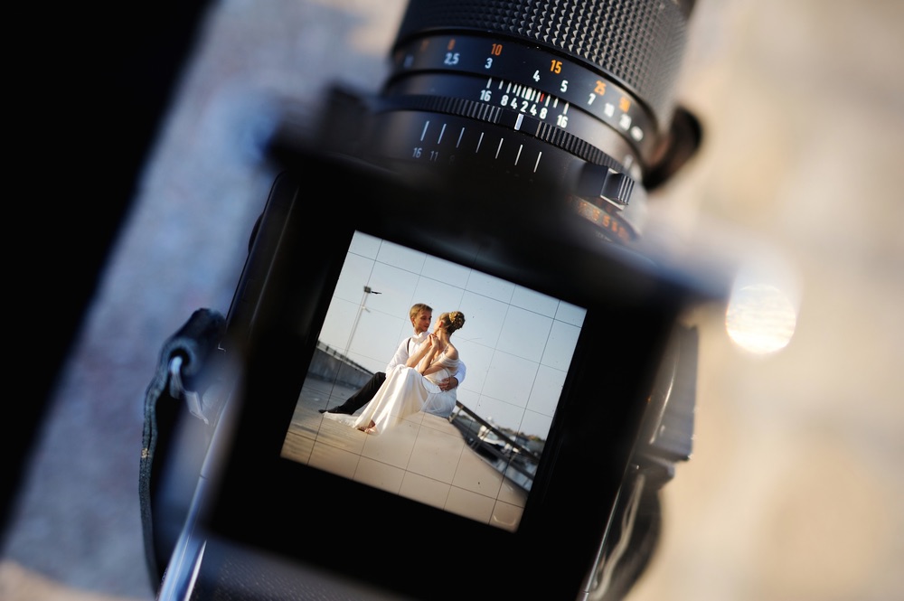 A photo looking through the electronic viewfinder of a camera at a bride and groom sitting on a ledge looking into each others eyes.