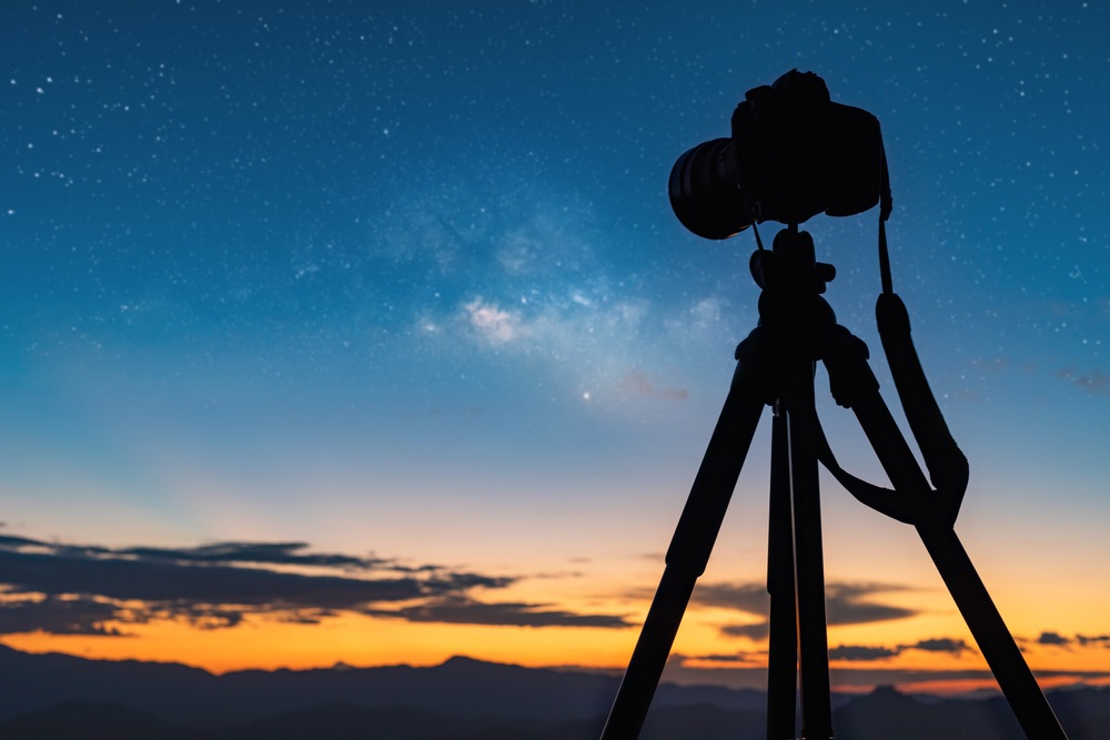 A camera resting on a sturdy tripod at night. Skyward view, showcasing the milky way.
