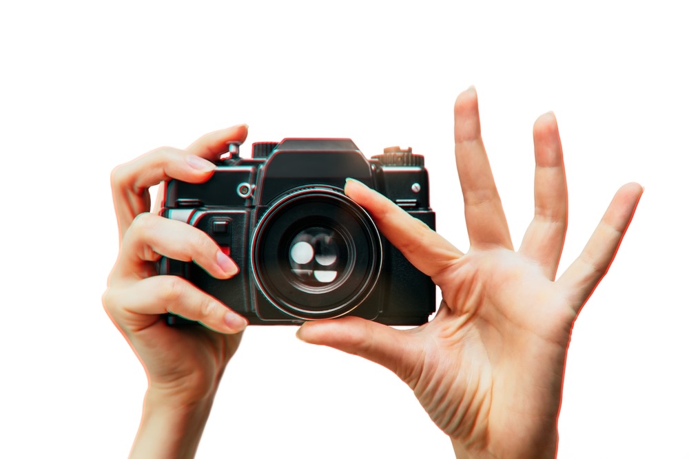A pair of hands is holding up a camera with the lens facing the viewer. One hand is holding the camera as if to push the shutter while the other is adjusting the lens focus.