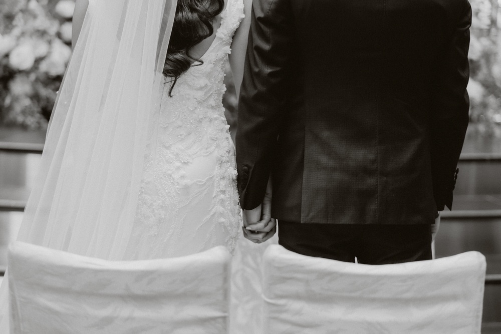 This is a candid black and white image of a bride and groom facing away from the photographer in front of some chairs holding hands. 