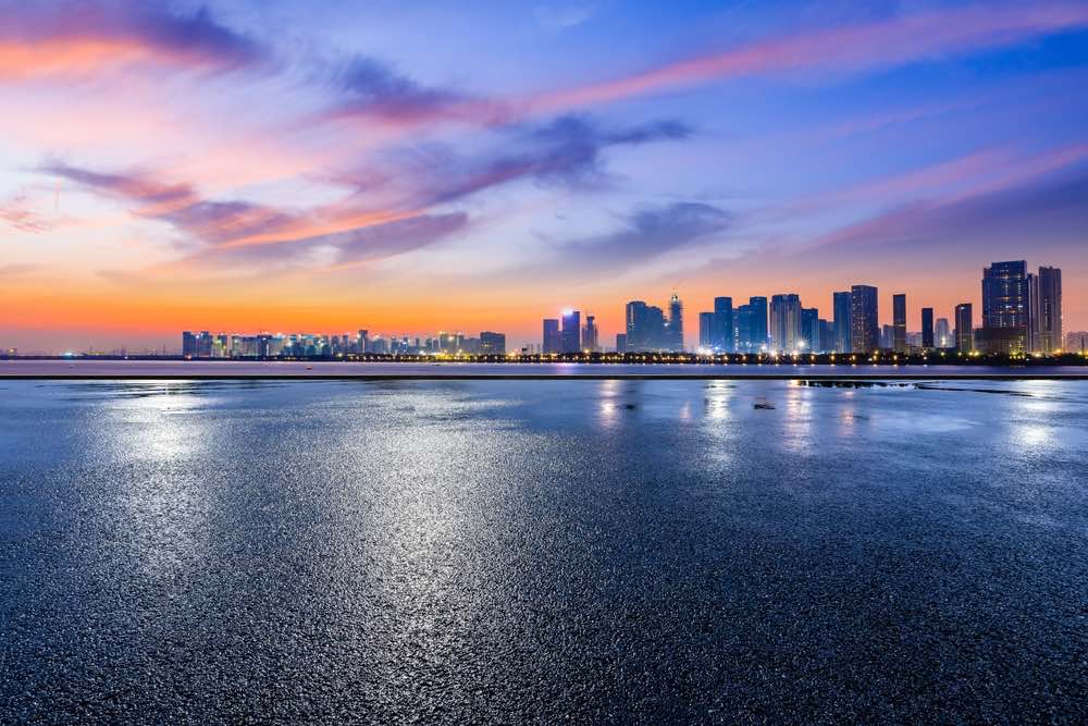 A sunrise photo from a low angle on some pavement overlooking a cityscape from across a body of water. The sky has orange, blue, and purple and street lights have come on.