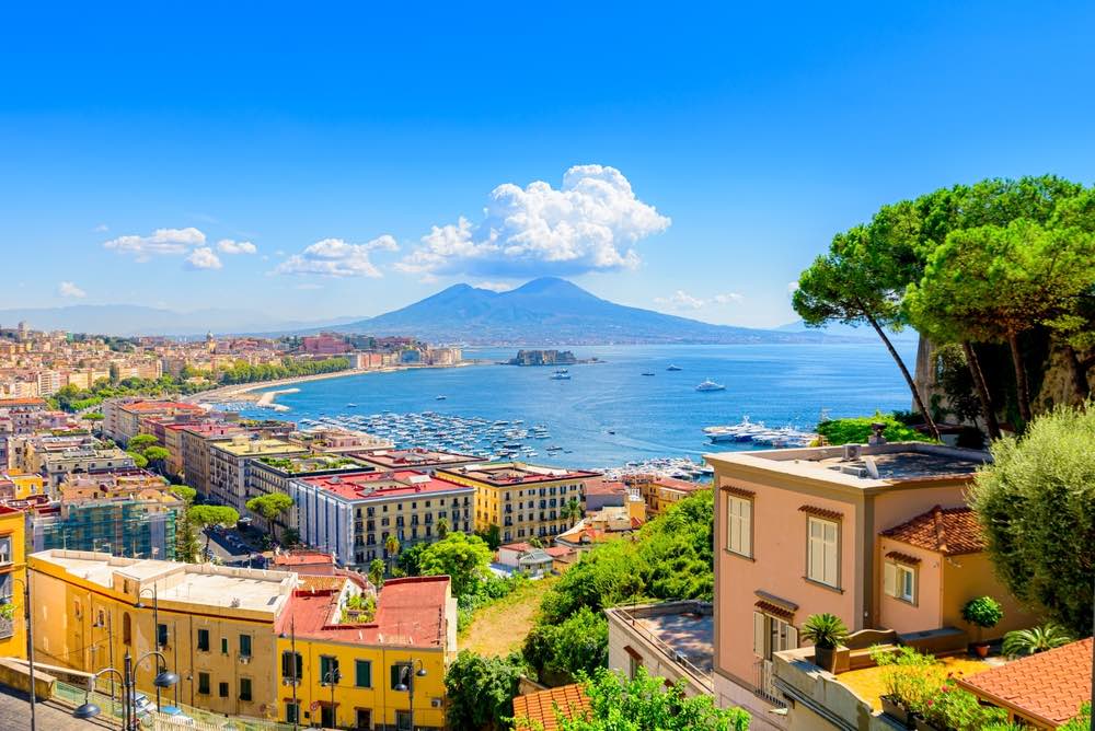 A view Mount Vesuvius in the background. The Mediterranean sea dominates the middle ground whilst the cityscape and foliage round out the foreground.