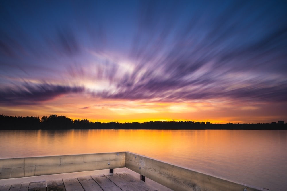 A photo during golden hours off a dock that is overlooking a body of water. It's a long exposure shot so the clouds are smooth and silky.