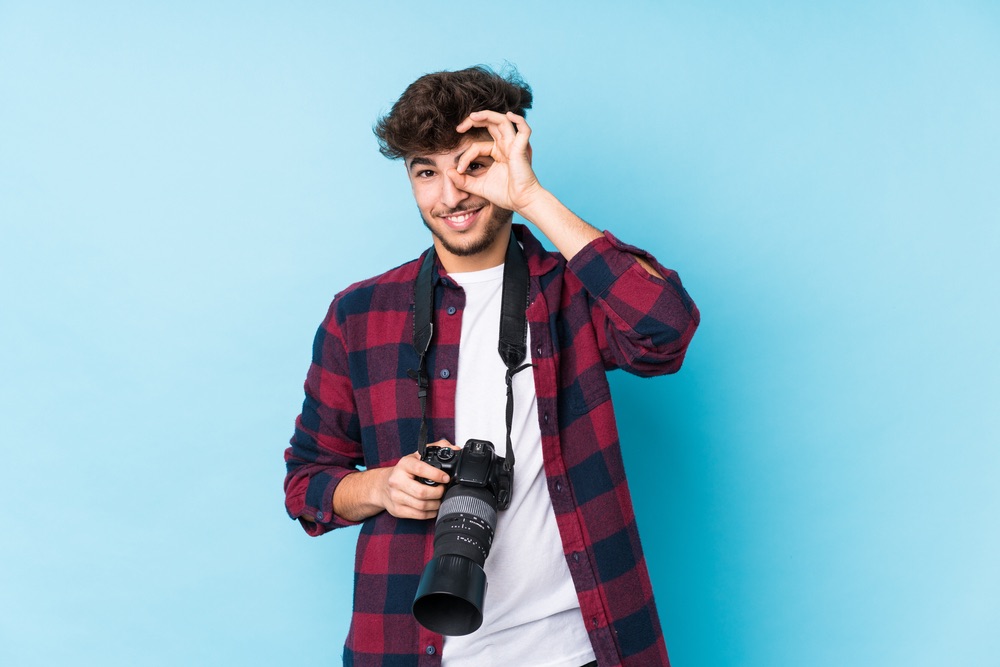 Portrait of a young male photographer gesturing 'okay' with his left hand. DSLR camera hanging from neck strap with zoom lens attached.