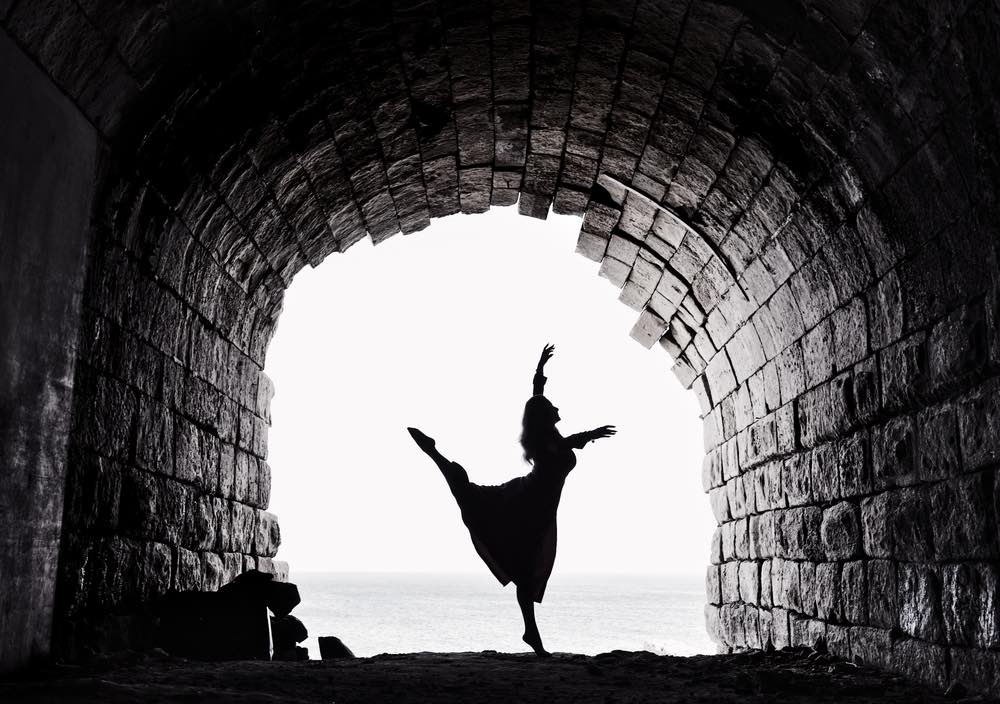 A dancer performing below an archway. Sea visible in the background. A black-and-white photo making great use of contrasts to build layers.