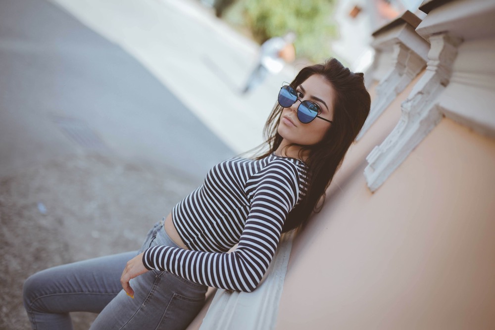 A dutch angle image of a young woman leaning up against the wall of a building. She has one leg cocked out, hand on her hip, and sunglasses halfway down her face.