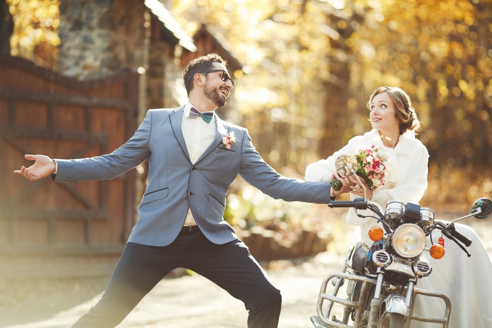 A wedding couple in a photo. The groom is extending some flowers to the bride while she sits on an old motorcycle.   