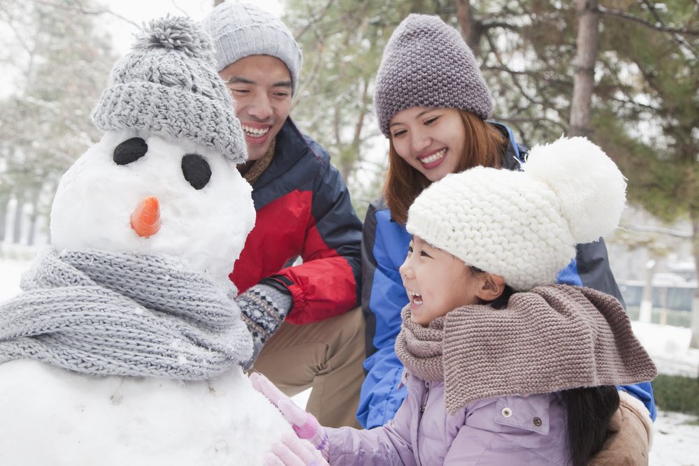 A mother, father, and young daughter build a snowman together in the snow.