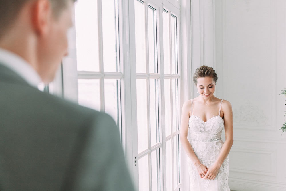 An image shot behind the groom that focuses on the bride accented by the natural lighting in the window.