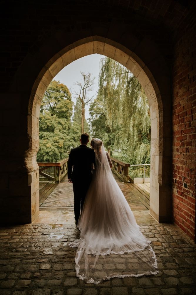 This image is of the backs of a bride and groom under an archway and surrounded by brick walls.