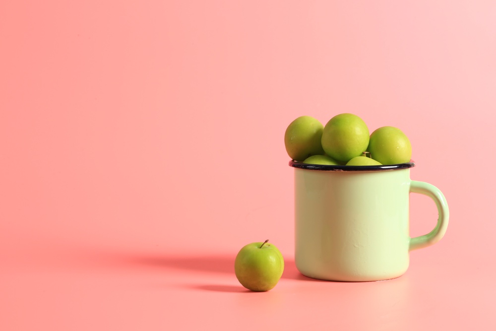 A mug full of apples sits on a pink surface. One of the apples is sitting upright outside of the cup.