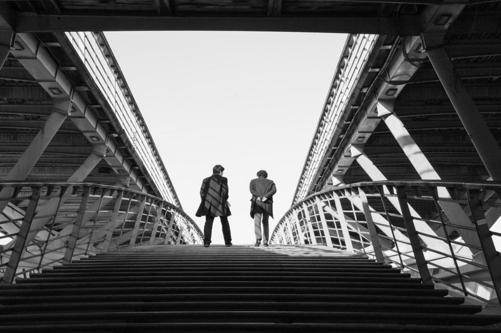 Two men ascend a staircase on a large footbridge. Black-and-white photograph.