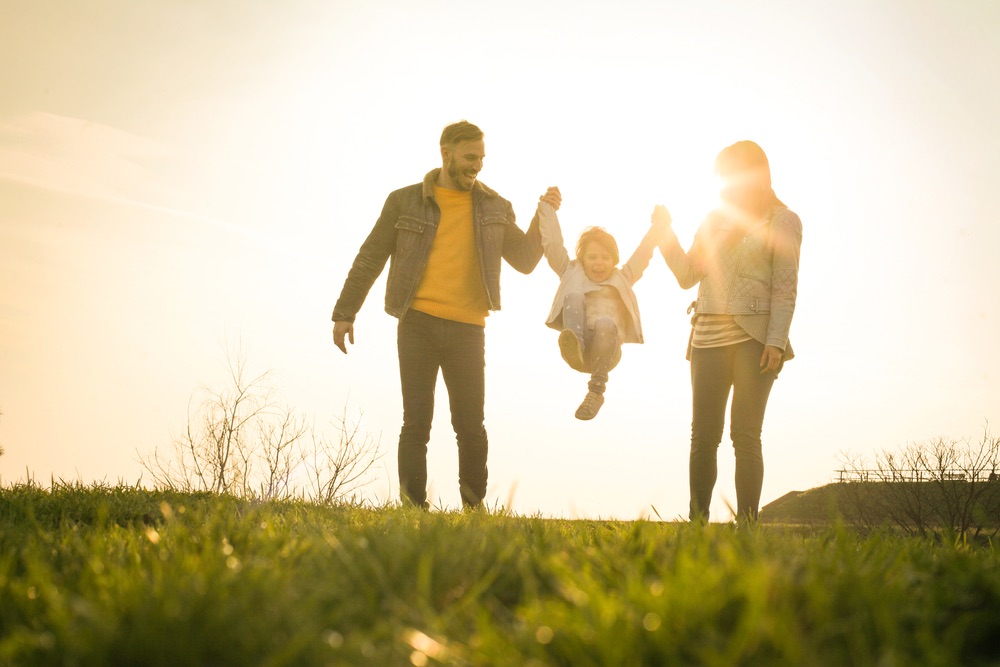 A father and mother are each holding a childs arm as they lift him in the air. The sun is behind them and they are on a grassy field. 