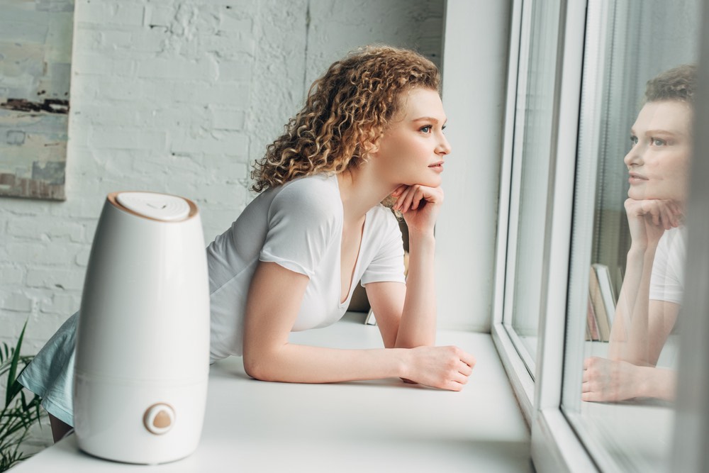 High-key scene of a young woman leaning over a windowsill. A small air purifier sits in the foreground.