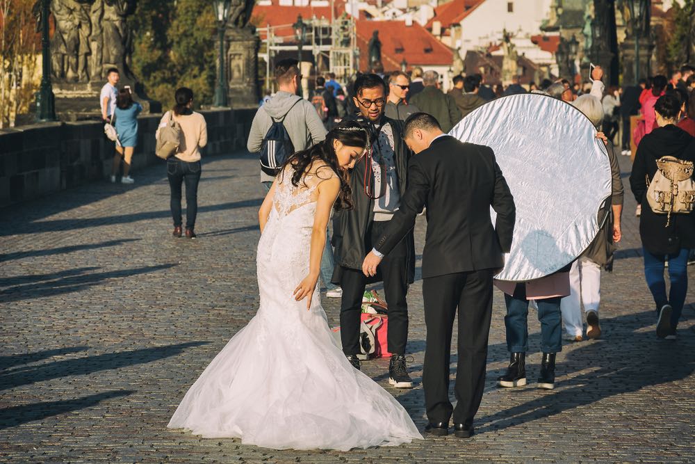 A wedding photographer and his assistant use a reflector to direct the sun light at the bride and groom while they take a photo.
