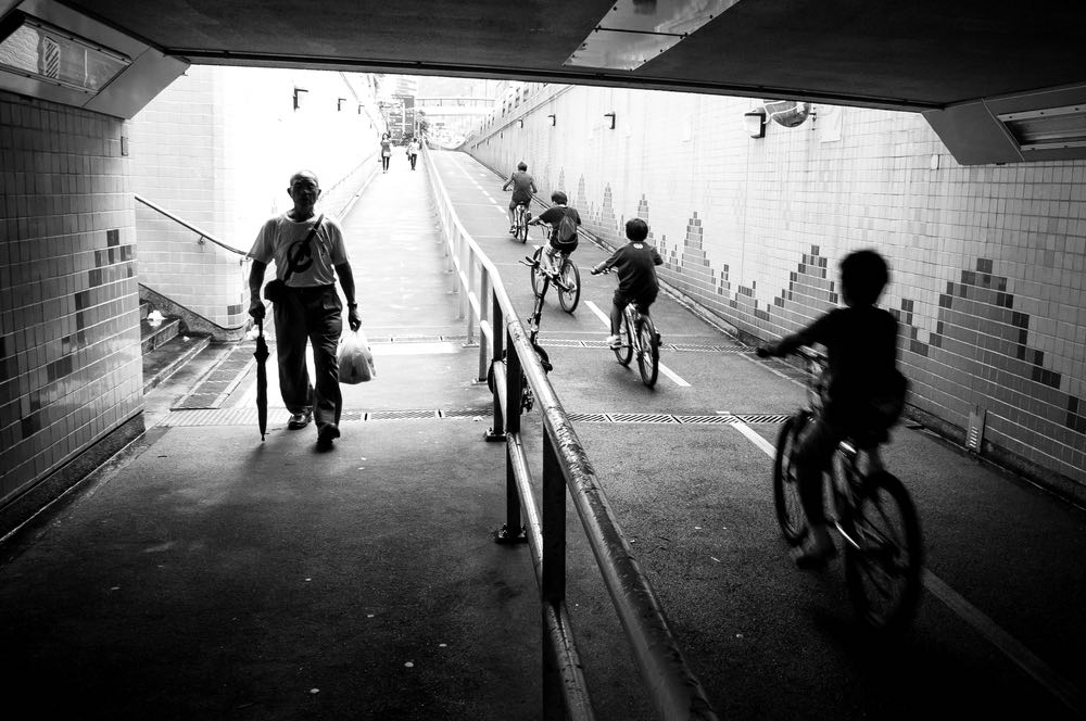 A group of children cycling through an underpass as older pedestrians walk by. Black-and-white street photography showcasing 'Bouncing Eye' composition.