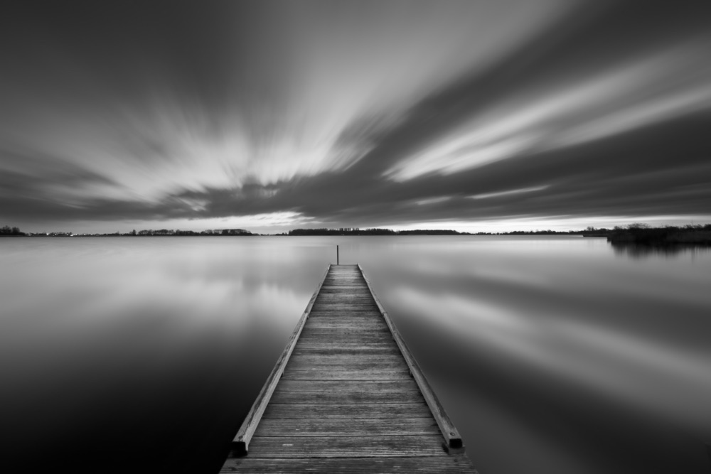 A black and white image from the shoreline looking down a dock towards a lake. It's a long exposure shot so the clouds and water are smooth.  