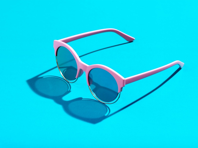 A pair of pink rimmed sunglasses are sitting on a turquoise surface. There is a light shining behind it which is cause the lens to cast a shadow in front of it.