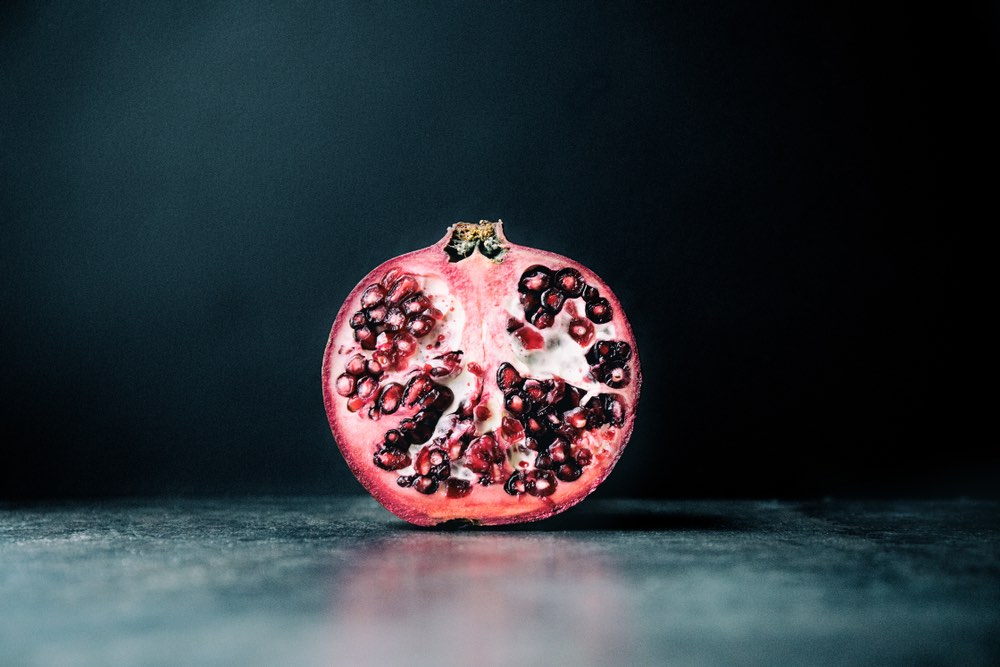 A pomegranate that has been sliced in half is sitting on a marble-like surface with a light off screen that is shining soft light onto it.