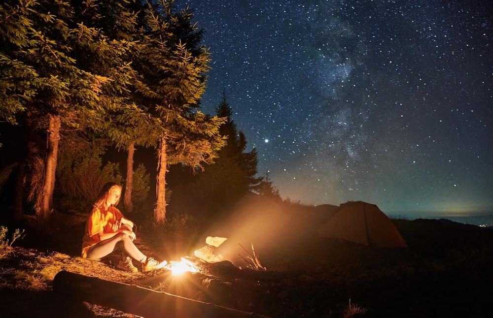 Single woman camping by fire in the forest. Starry sky with milky way visible at night. Tent in background.