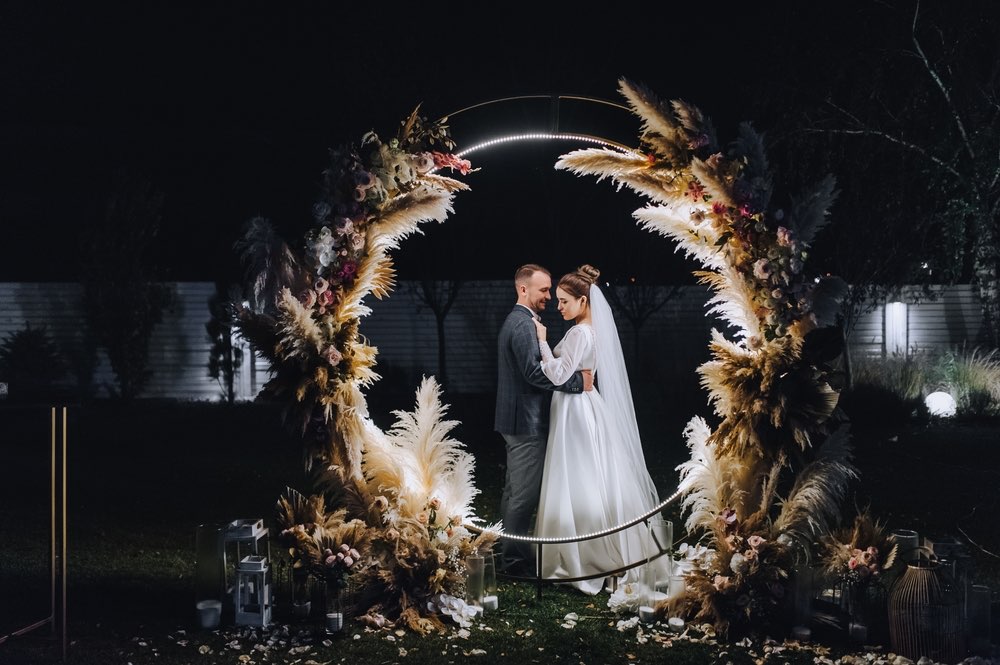 A wedding couple inside a decorated wreath that is illuminated with lights during a nighttime photography shoot.