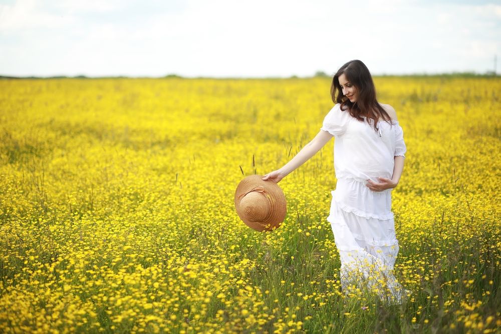 An expectant mother in a white dress poses out in the middle of some farm pasture.