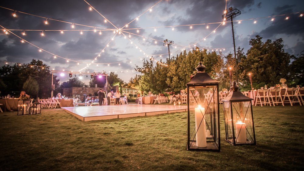 An outdoor wedding on an overcast day. There are lights overhead and candles in the foreground. The dancefloor is surrounded by chairs. 