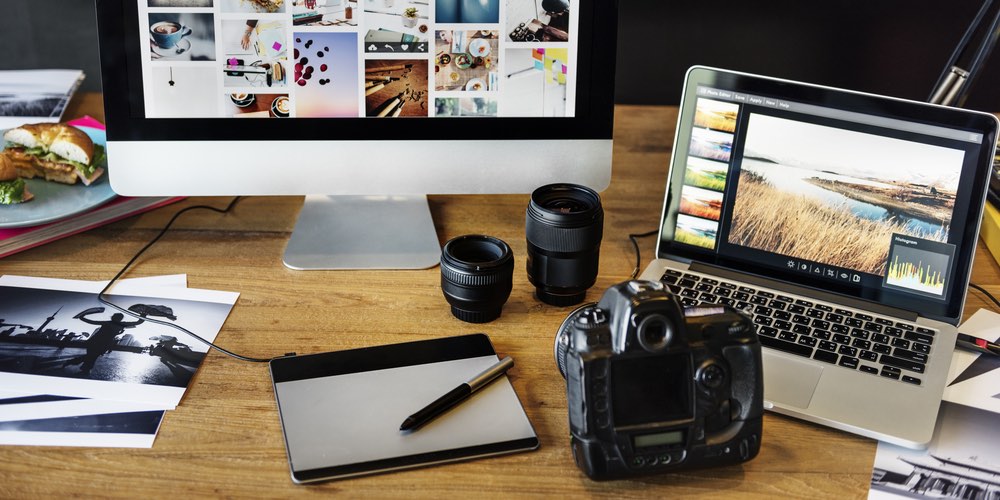 An example of a photo editing workstation, showing an array of common digital photography post-processing gear. DSLR camera, stylus pen and drawing tablet, laptop and desktop computer as well as prints visible.