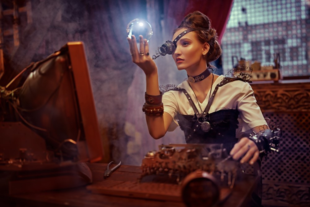 A woman in steampunk style attire holds a crystal ball up to her face which is illuminated.