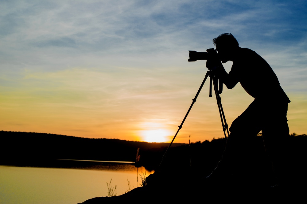 A silhouette of a photographer taking a picture with his camera on a tripod with a zoom lens. The sun is setting from beside him.