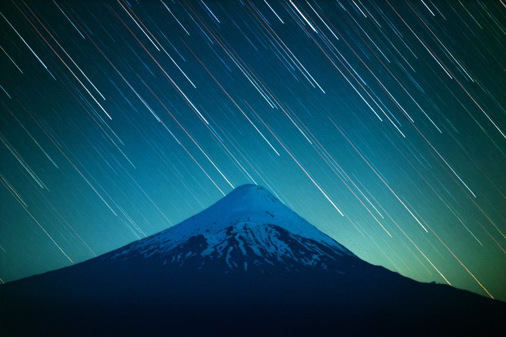 View of a mountaintop in blue with star trails in the background. Captured using a single-exposure method instead of image stacking, leaving noticeable "shooting star"-like trails without gaps.