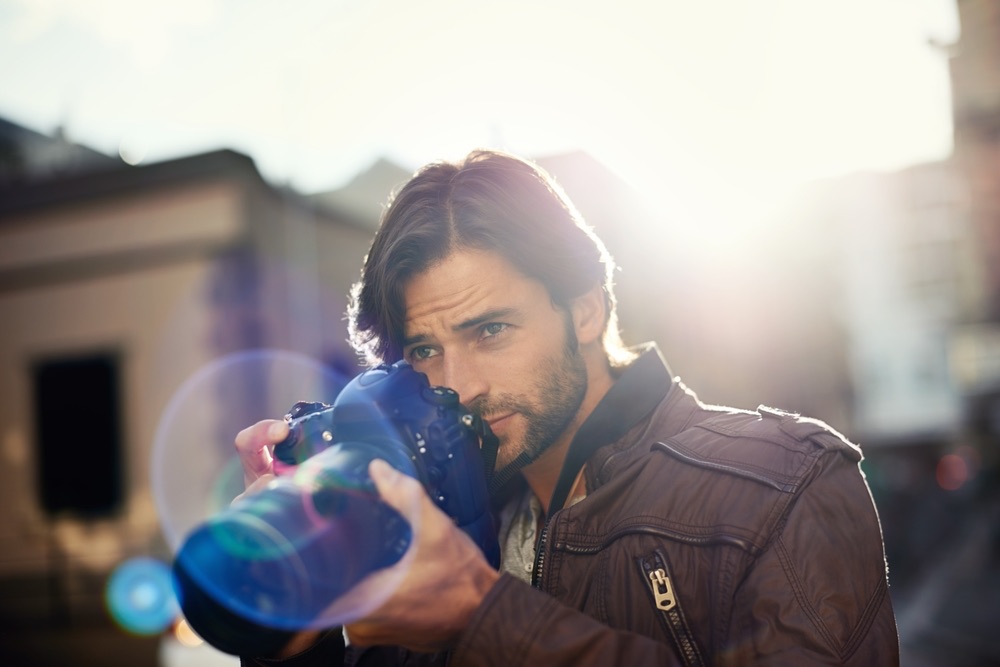 A young male photographer composing a shot in the street. Bloom and lens flare effects visible in background.