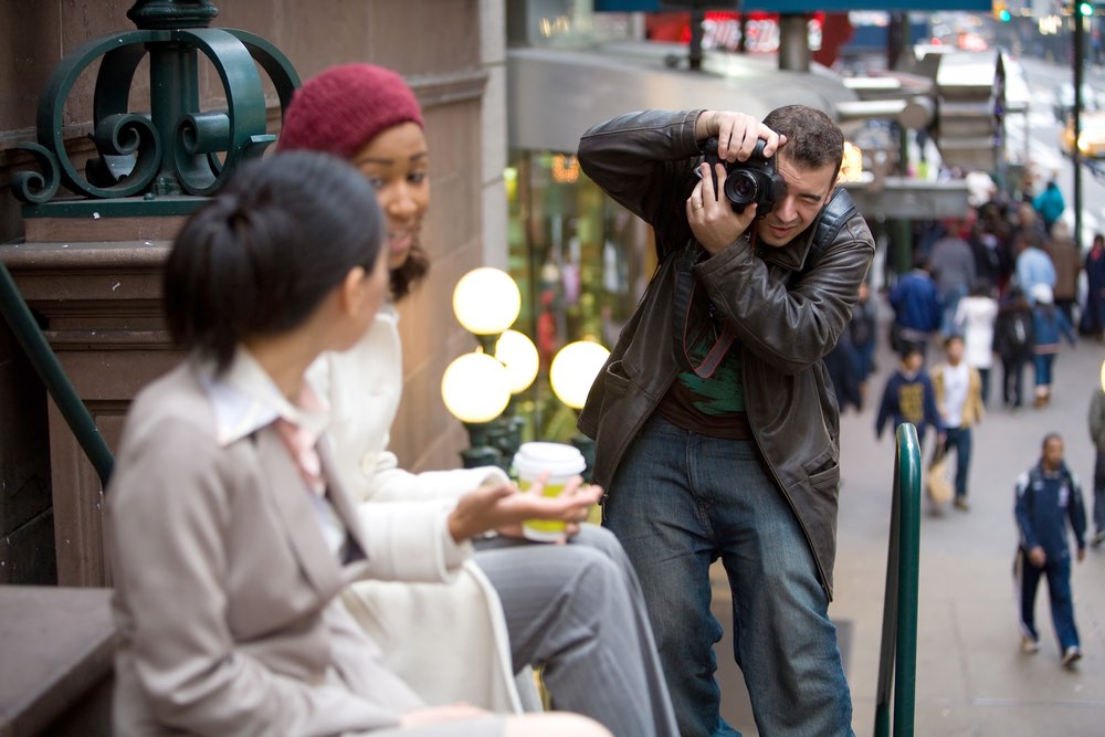 A street photographer taking pictures of two young women in a public environment. Consent is an essential component of ethical photography conduct in situations like this.