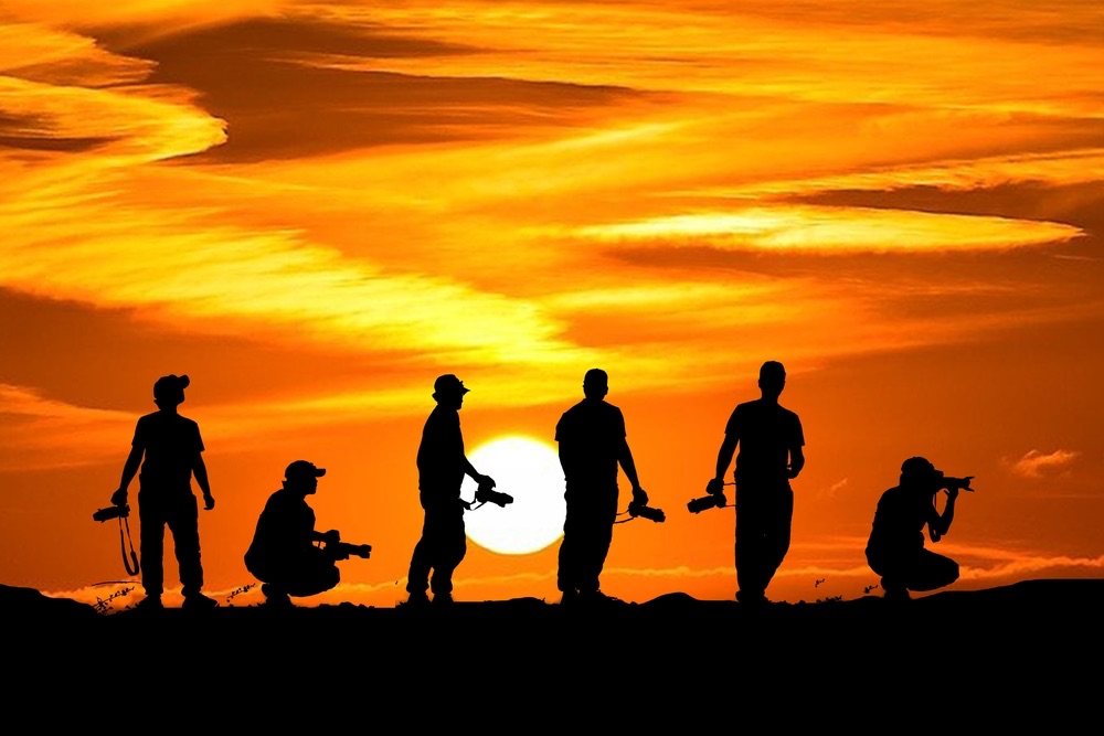 A group of photographers posing at sunset. Orange sky background with silhouettes of photographers outlined in black.