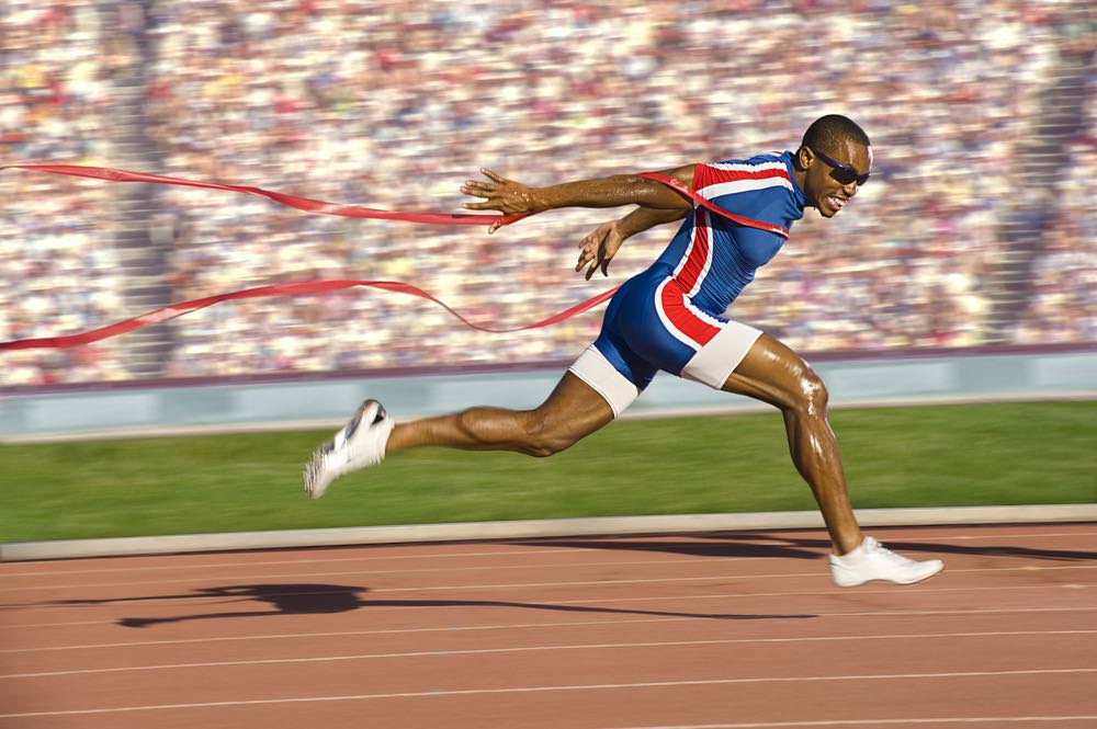 A man is sprinting across the finish line in a running race. There is a track underneath him and a blurry crowd in the background.  