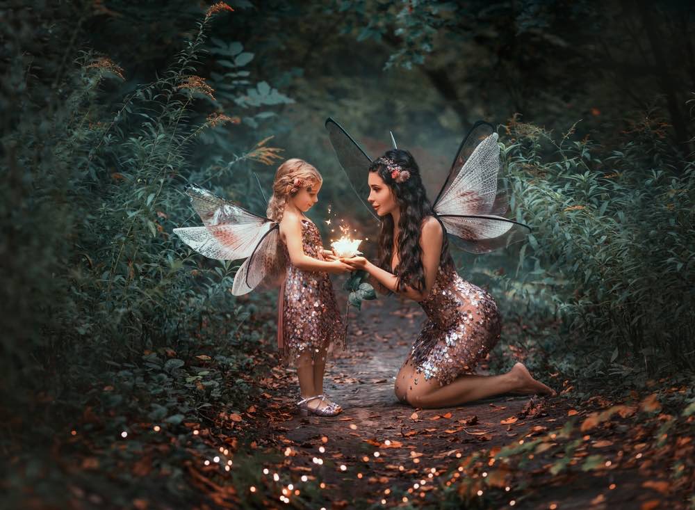 Two female fairies are holding a glowing flower together in the middle of a forest path for fantasy photography.