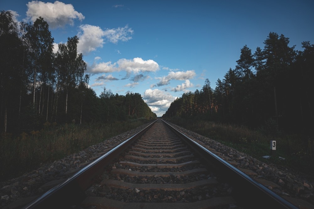 A shot of a railroad track with forest lining both sides of the frame. An underexposed image, with a clear sky but poorly-defined shadows and murky details in darker areas.