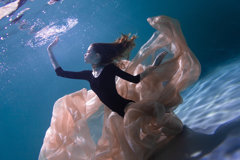 A woman in a flowing tutu is underwater touching the surface of the water from below.