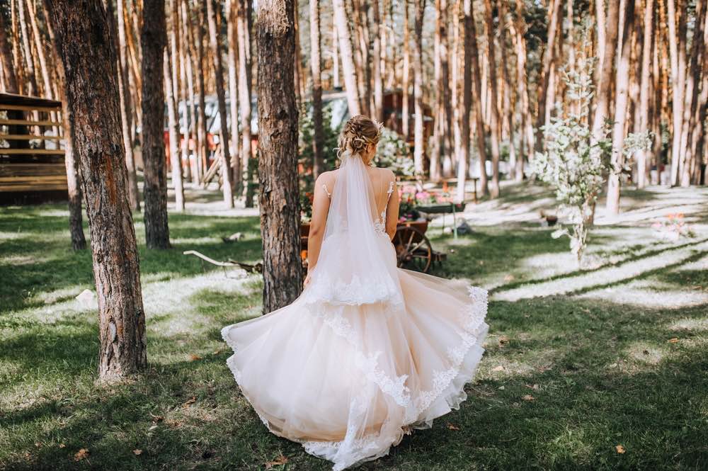 A blonde bride is walking into a forest that is lit with dappled sunlight. 