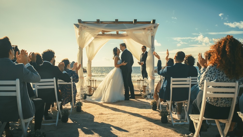 A photo of an outdoor wedding ceremony near water shooting up the aisle at the bride and groom. Chairs on either side filled with people.
