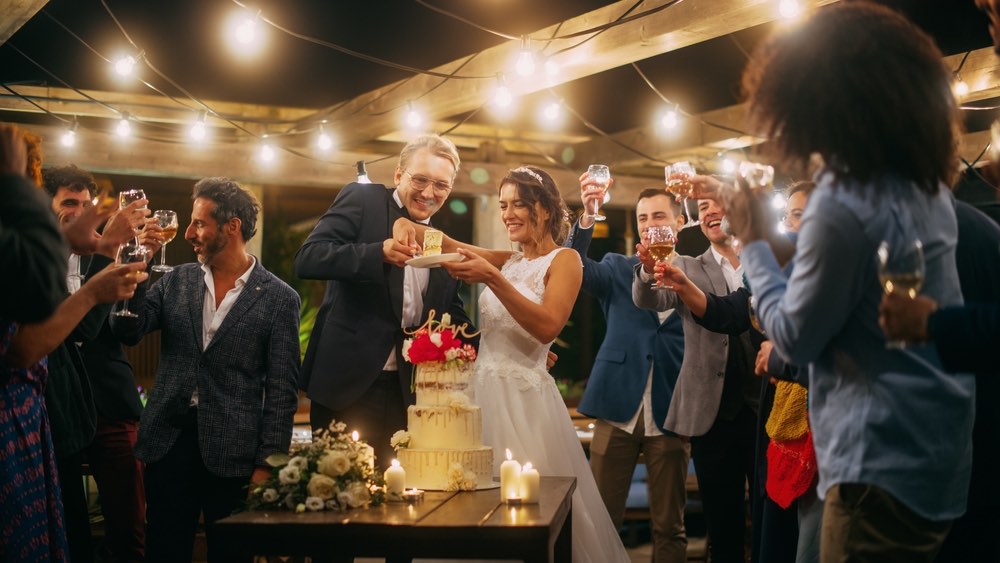 A well-lit outdoor wedding reception with the bride and groom cutting the cake surrounded by wedding guests. 