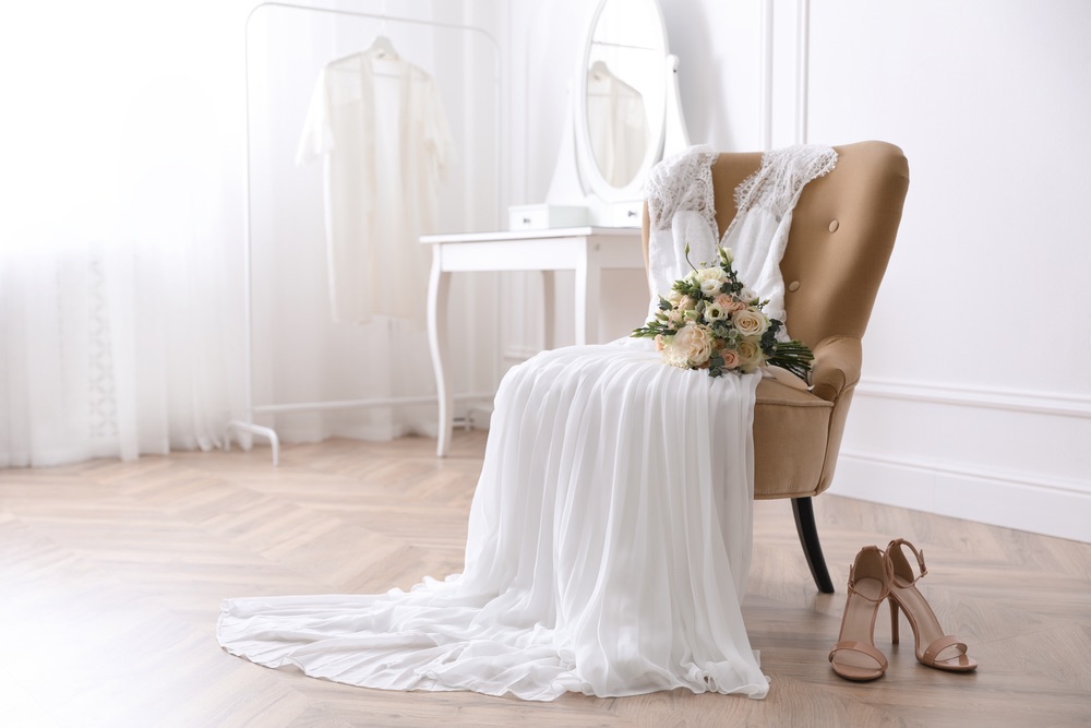 An image of a wedding dress draped over a chair. A bouquet is seated on the dress and the high heels are set beside the chair. 