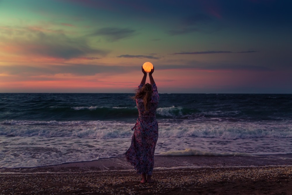 Photograph of a woman standing on the beach, holding the Moon above her head in her outstretched hands.