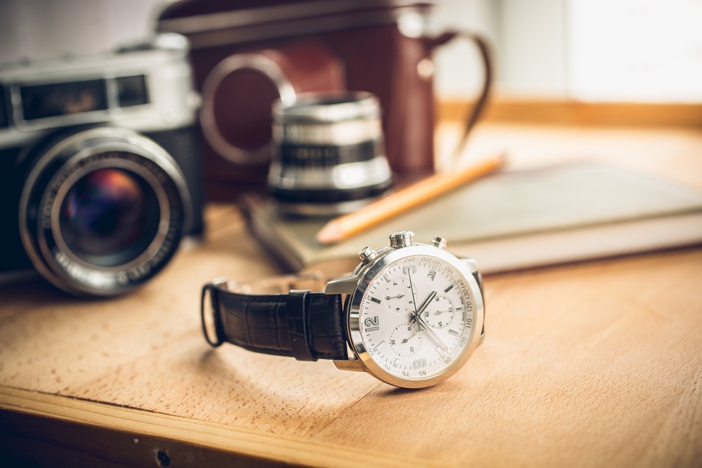 A close-up shot of a wristwatch with some photographic equipment sat behind on a desk. Wide-aperture shot showcasing some bokeh.
